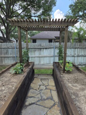 Another angle of two parallel cedar raised garden beds connected with an arbor trellis finished with a flagstone path, a sitting bench, and a few plants planted around the trellis.
